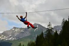 Mann winkt beim Abseilen auf einer Seilbahn mit Berglandschaft im Hintergrund